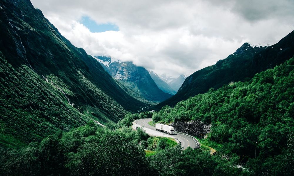 The image depicts a winding mountain road cutting through a breathtaking valley surrounded by towering, snow-capped peaks. The road twists and turns, leading the eye deep into the rugged landscape. A few large trucks can be seen navigating the curves, their presence dwarfed by the immense scale of the natural environment. The green vegetation clings to the steep slopes, contrasting with the rocky formations and adding vibrant color to the scene. Clouds hang low, partially obscuring the distant mountain tops and adding a sense of atmosphere and drama to the composition.