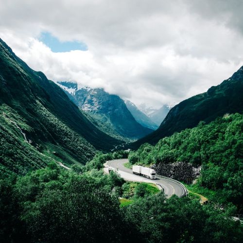 The image depicts a winding mountain road cutting through a breathtaking valley surrounded by towering, snow-capped peaks. The road twists and turns, leading the eye deep into the rugged landscape. A few large trucks can be seen navigating the curves, their presence dwarfed by the immense scale of the natural environment. The green vegetation clings to the steep slopes, contrasting with the rocky formations and adding vibrant color to the scene. Clouds hang low, partially obscuring the distant mountain tops and adding a sense of atmosphere and drama to the composition.